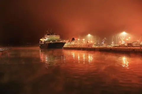 A ship alongside a quay in fog, the lights turning the air orange