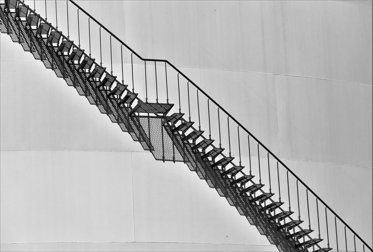 A steel staircase winding up the side of a white storage tank, in black and white