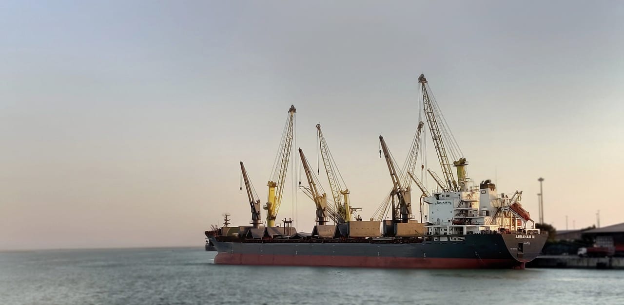 A bulk carrier with its deck cranes raised, off a hazy coast