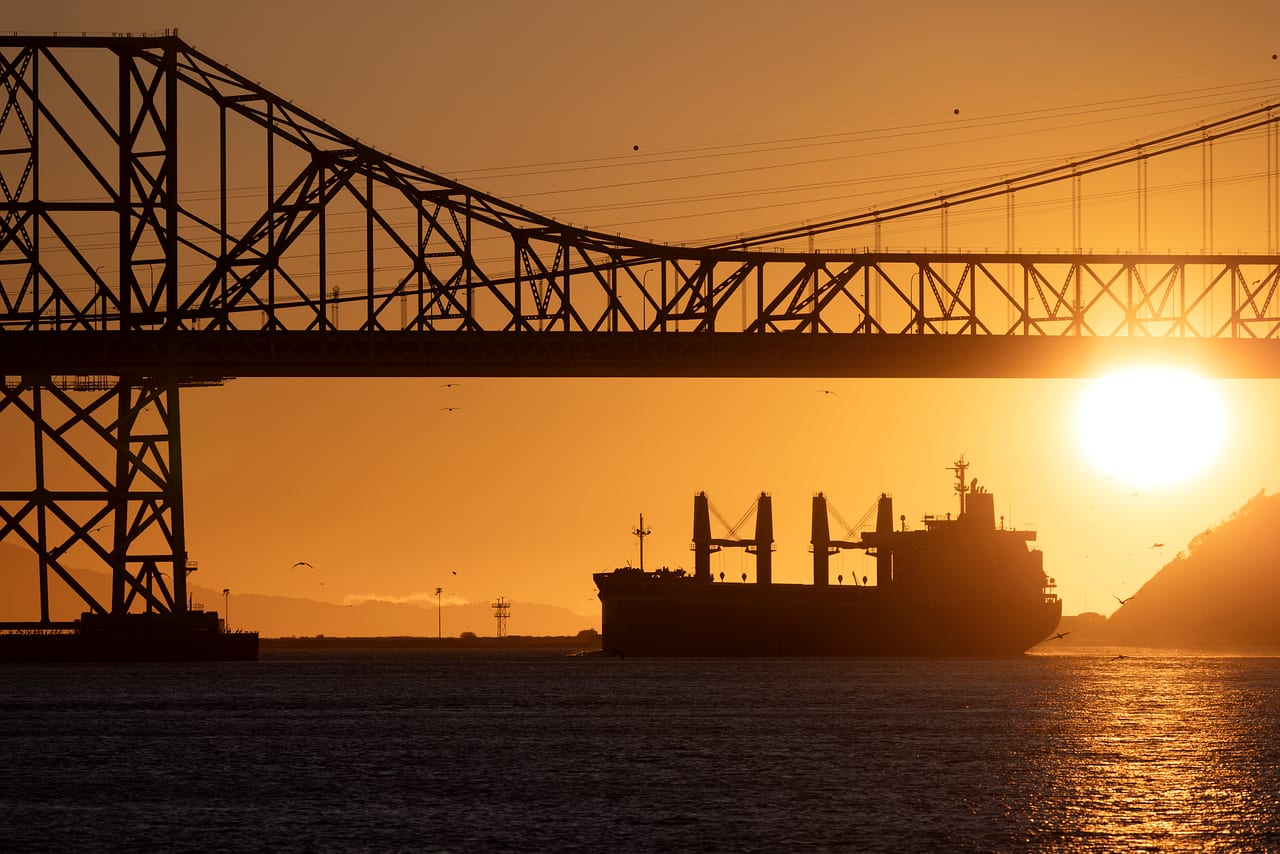 A bulk carrier in silhouette passing under a bridge at sunset